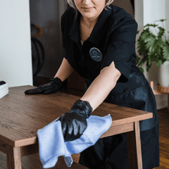 A woman wearing black gloves and a black uniform cleaning a wooden table with a purple cloth in a well-lit room.