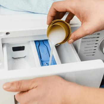 A close-up of hands pouring liquid detergent into a washing machine drawer with detergent powder already inside.