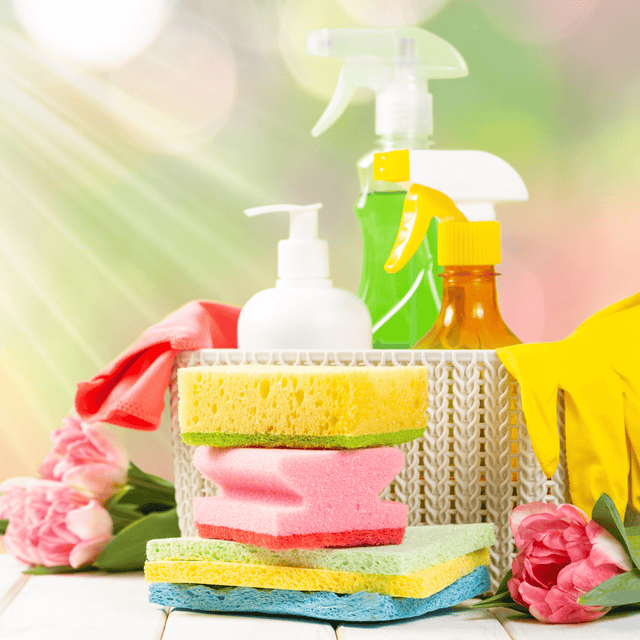 A collection of colourful cleaning supplies including sponges, spray bottles, gloves and a soap dispenser, set outdoors with flowers and sunlight in the background.