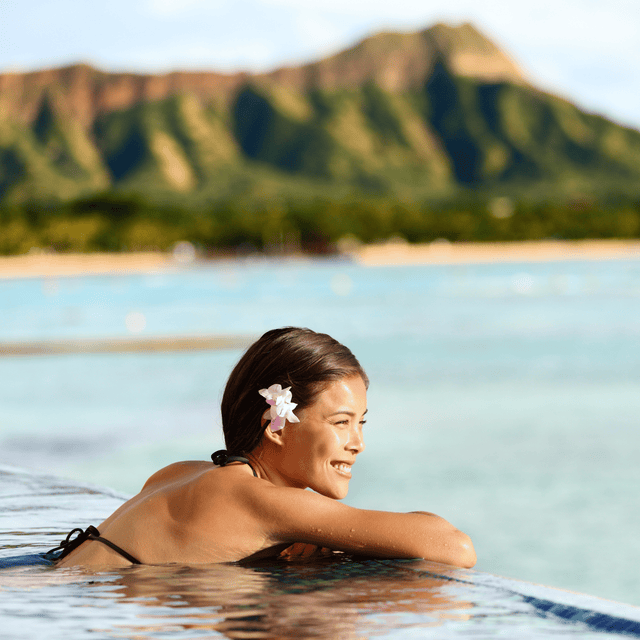 A woman with a flower in her hair relaxing in an infinity pool with a scenic view of the ocean and lush green mountains in the distance.