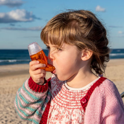 Child using AirPhysio for Children breathing device at the beach, gently exhaling through the mouthpiece to support airway clearance